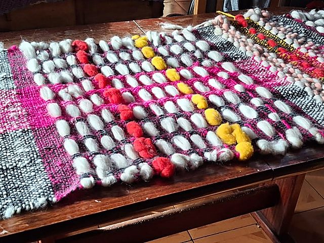 Piecera tejida por Sandra Zurita, con cuadros grises y fucsia y pompones de colores